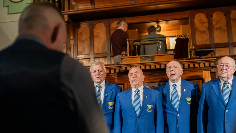 Older men in matching blue blazers singing beside an organist